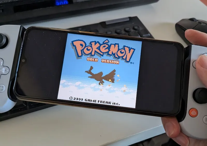 Gameboy game Pokemon Gold on a Samsung A23 phone. The screen shows the pokemon Ho&#x27;oh flying in a blue sky. The pokemon resembles a phoenix. This is in a pixilated style.